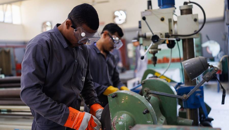 عاملان داخل ورشة يرتديان نظارات واقية وقفازات، ويعملان على آلة جلخ المعادن | تعبيرية