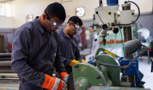 عاملان داخل ورشة يرتديان نظارات واقية وقفازات، ويعملان على آلة جلخ المعادن | تعبيرية