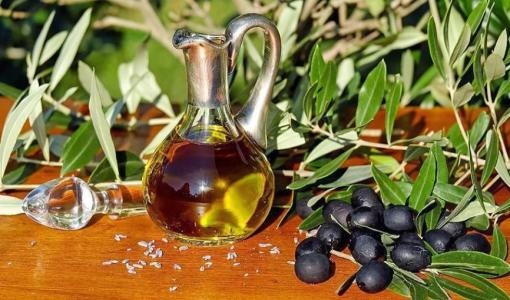 إبريق زجاجي مليء بزيت الزيتون وإلى جانبه ثمار زيتون سوداء وأغصان زيتون خضراء