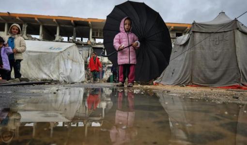 طفلة غزية تحمل مظلة أكبر من حجمها، تحاول الاحتماء بها من المطر وتنعكس صورتها في بركة ماء أمامها، بالقرب من خيام مهترئة ومبانٍ مدمرة جراء حرب الإبادة الإسرائيلية في قطاع غزة | المصدر: TRT