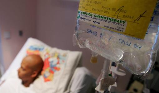 كيس علاج كيميائي معلق في غرفة مستشفى، بينما يبدو في الخلفية مريض سرطان مستلقٍ على السرير | Getty Images