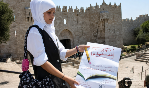 إحدى الطالبات تقف عند باب العامود في القدس المحتلة و تحمل منشورا توعويا يدعو لحماية المنهاج الفلسطيني من الأسرلة
