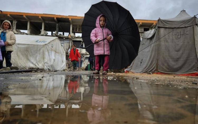 طفلة غزية تحمل مظلة أكبر من حجمها، تحاول الاحتماء بها من المطر وتنعكس صورتها في بركة ماء أمامها، بالقرب من خيام مهترئة ومبانٍ مدمرة جراء حرب الإبادة الإسرائيلية في قطاع غزة | المصدر: TRT