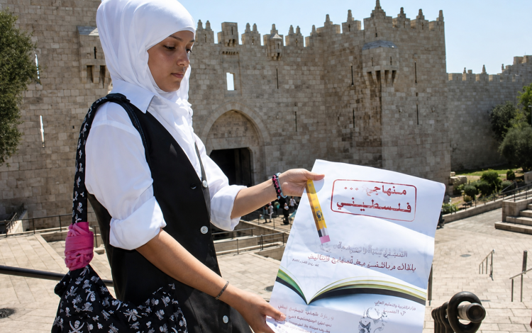 إحدى الطالبات تقف عند باب العامود في القدس المحتلة و تحمل منشورا توعويا يدعو لحماية المنهاج الفلسطيني من الأسرلة